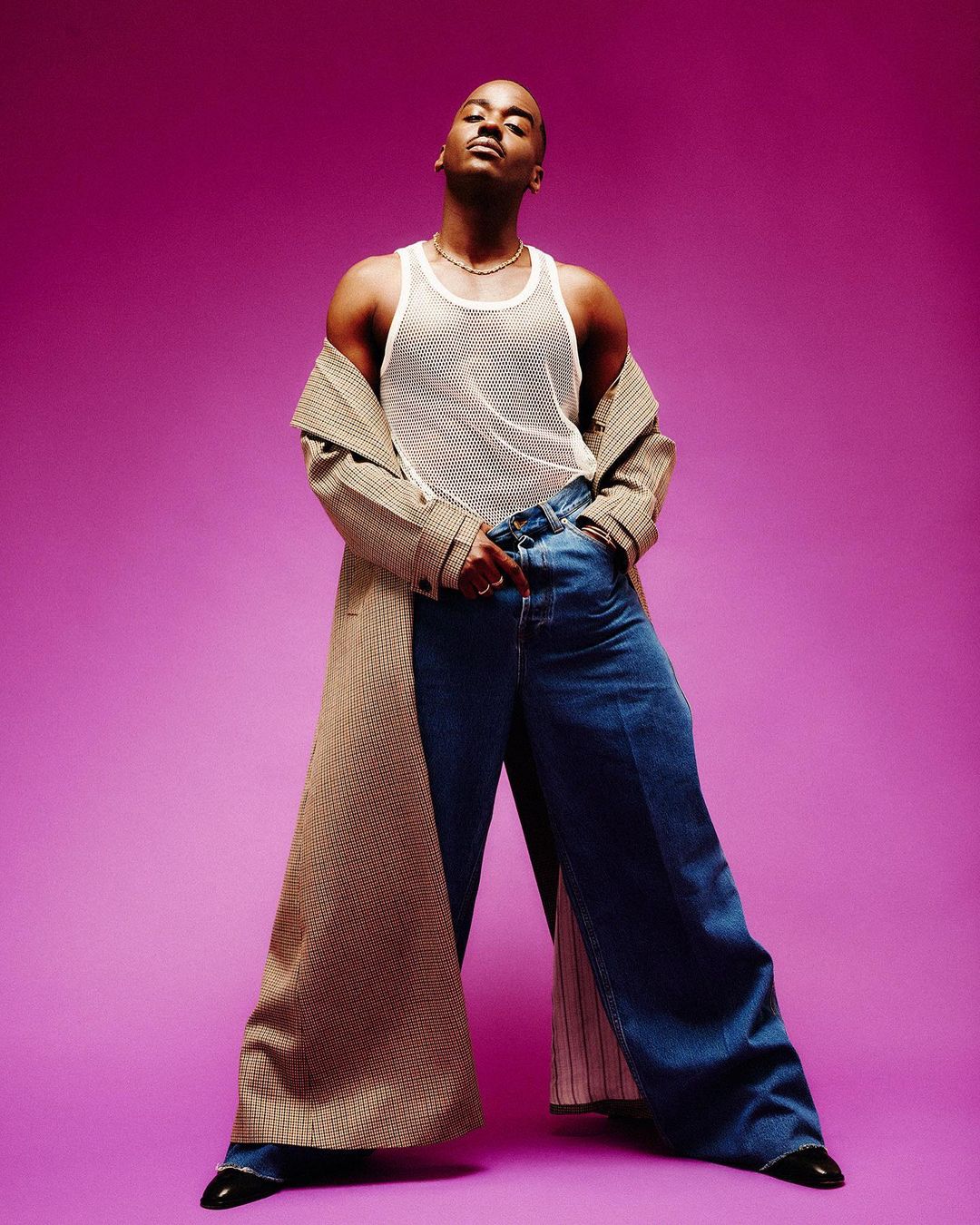 Stylish adult male poses in an open coat with wide-leg jeans against a vibrant purple background, showcasing a blend of modern fashion and casual elegance.
