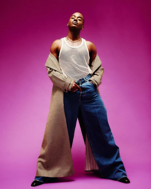 Stylish adult male poses in an open coat with wide-leg jeans against a vibrant purple background, showcasing a blend of modern fashion and casual elegance.