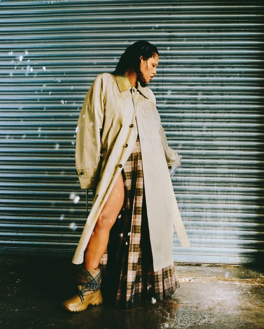 Woman poses in a beige trench coat over a plaid skirt, paired with lug-soled boots, against a corrugated metal backdrop.