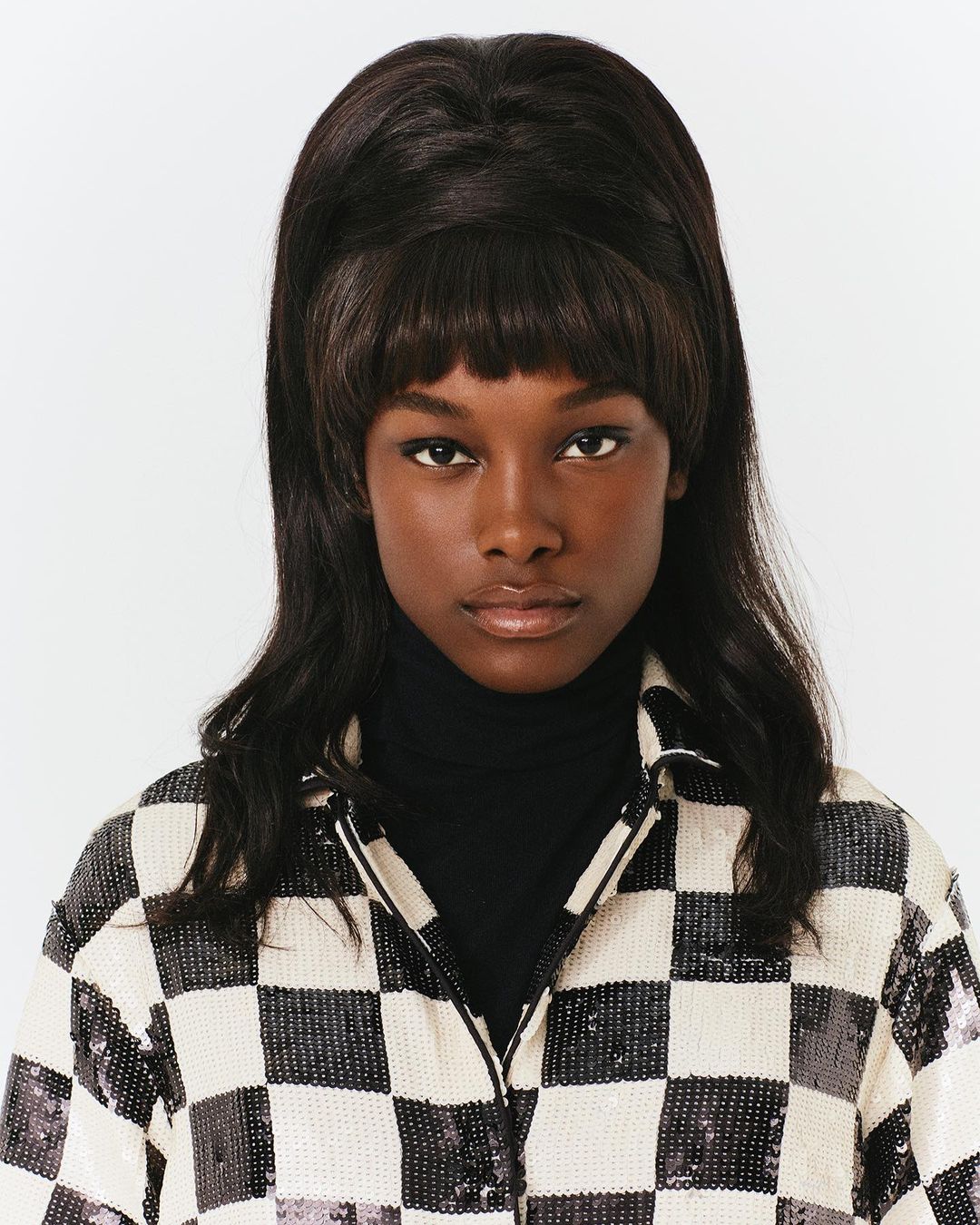 Close-up portrait of a Black woman with dark beehive hair and bangs wearing a black turtleneck and a white and black checkered jacket.