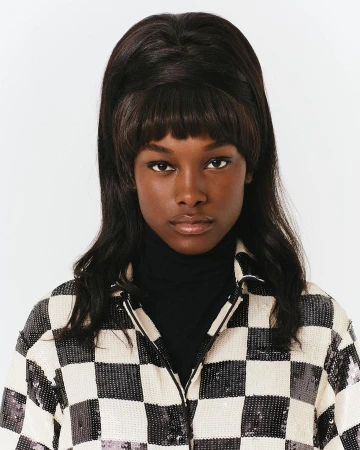 Close-up portrait of a Black woman with dark beehive hair and bangs wearing a black turtleneck and a white and black checkered jacket.