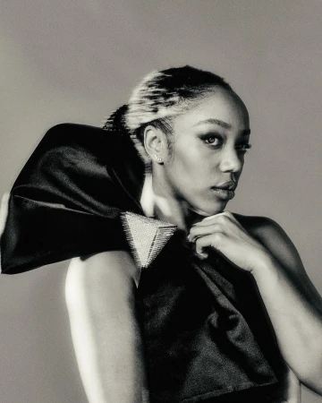 Monochromatic studio portrait of an adult woman with fair skin and dark hair styled back, wearing a black satin garment with embellishment.