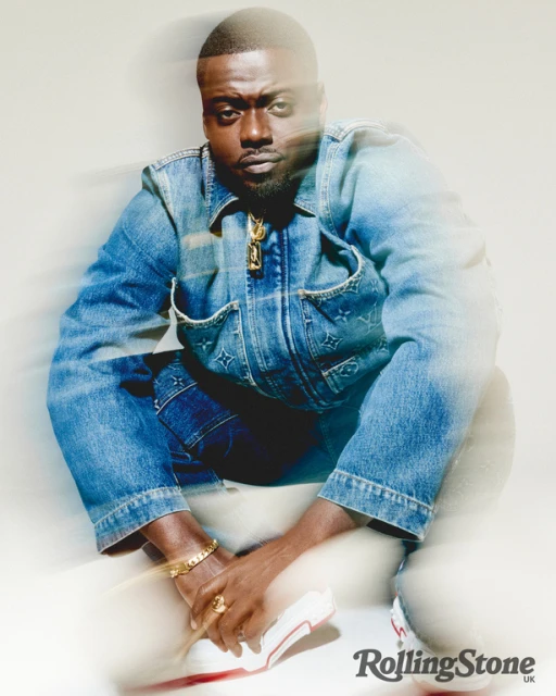 Portrait of Daniel Kaluuya squatting and looking at the camera, wearing a denim jacket and pants, with a gold necklace.