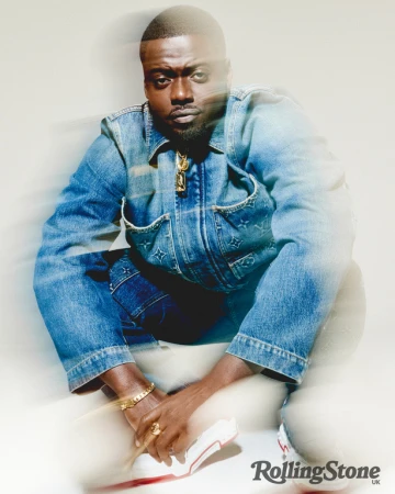 Portrait of Daniel Kaluuya squatting and looking at the camera, wearing a denim jacket and pants, with a gold necklace.