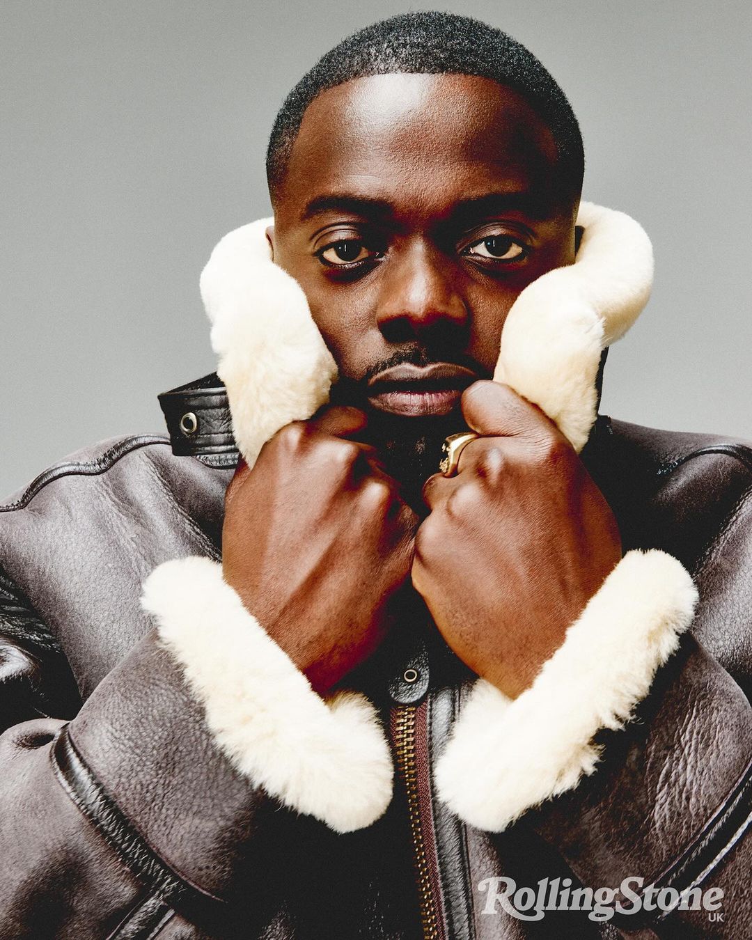 Close-up portrait of an adult male with dark hair, wearing a brown jacket with cream-colored shearling collar, against a neutral background.