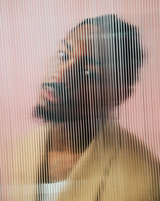 Close-up portrait of an adult Black male with a tan jacket, head tilted back, visible through reeded glass against a pale pink background.