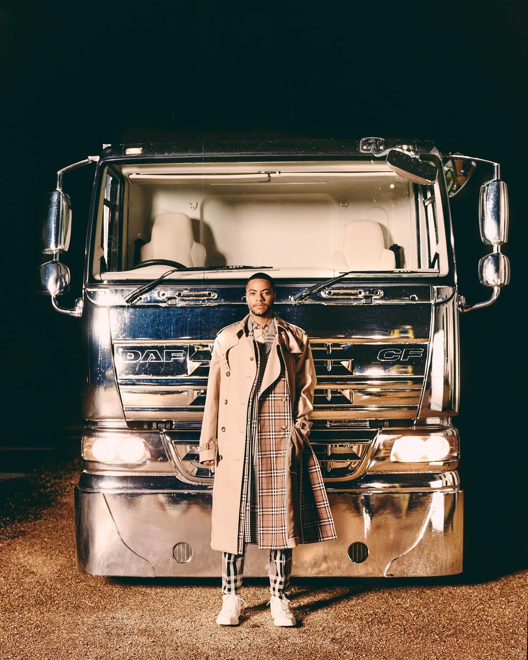 Adult male in a beige trench coat stands in front of a large, chrome-plated DAF CF truck with a dark, out-of-focus background.