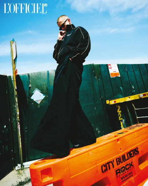 Stylish man in black track suit and sunglasses posing on orange barrier in urban setting beneath a partly cloudy blue sky.