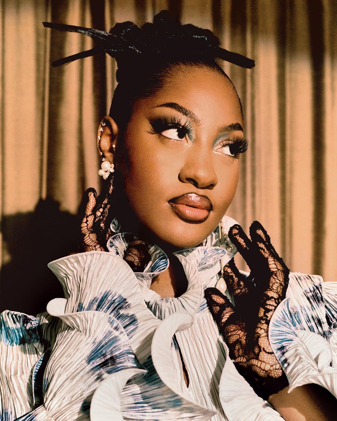 Portrait of a stylish adult female with dramatic makeup, wearing a pleated top, lace gloves, and hair sticks, against a draped backdrop.