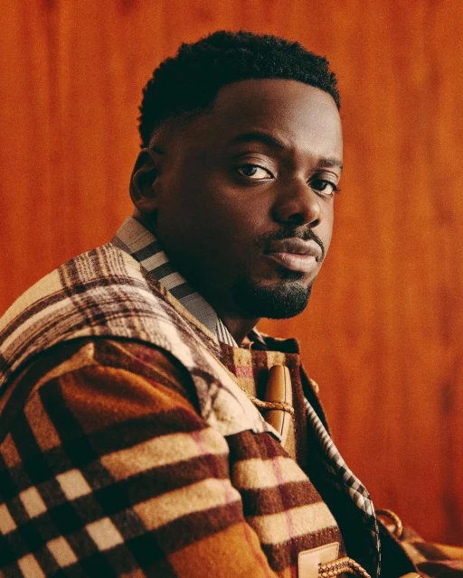 Portrait of adult male with short hair, wearing a brown, tan, and white plaid jacket looking into the camera against an orange wall.