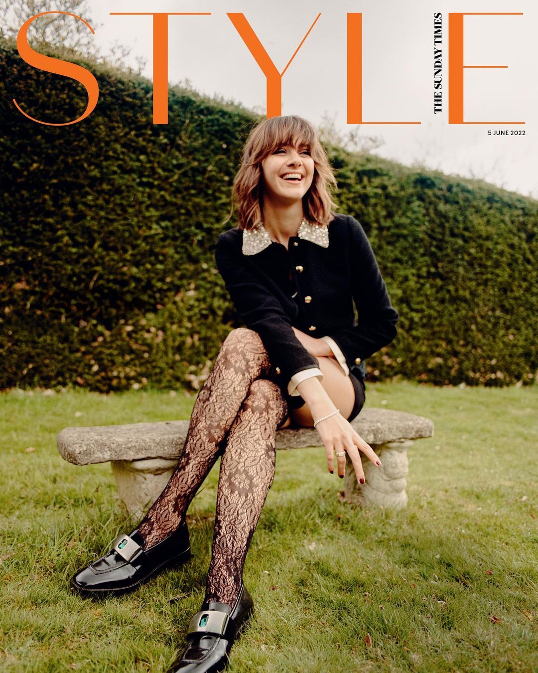 Smiling woman in dark jacket and patterned tights seated on stone bench outdoors against a clipped hedge, photographed by Danny Kasirye.
