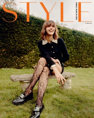 Smiling woman in dark jacket and patterned tights seated on stone bench outdoors against a clipped hedge, photographed by Danny Kasirye.