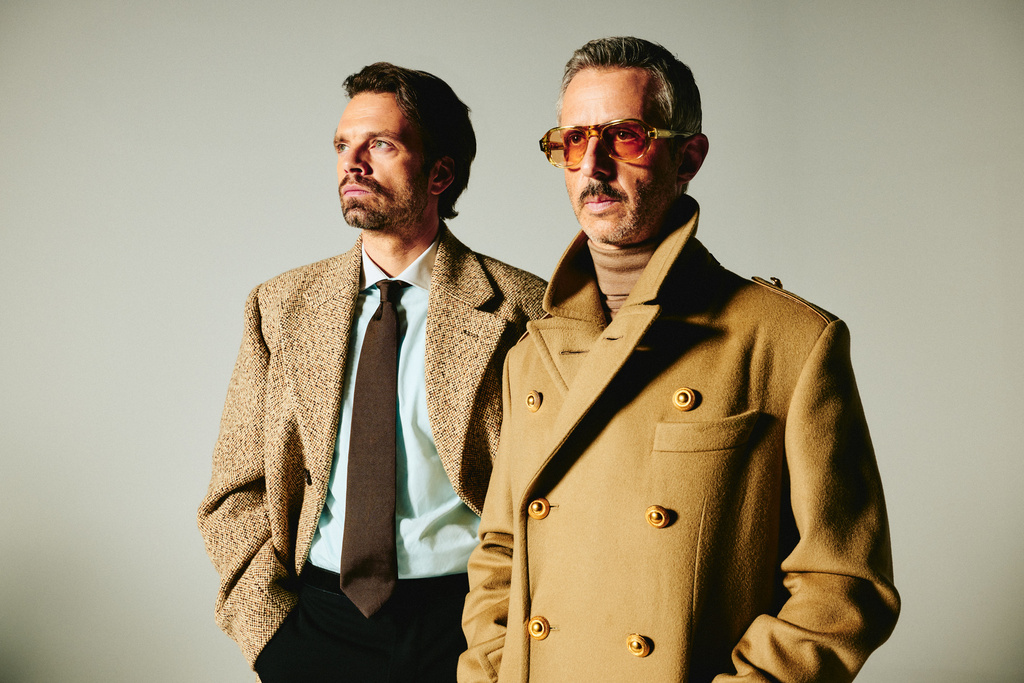 Two stylish adult males in neutral-toned coats and ties pose in a studio setting, showcasing fashion and editorial style.