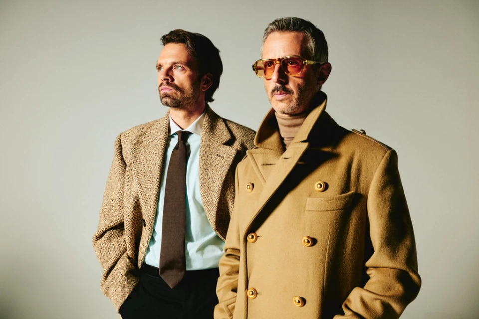 Two stylish adult males in neutral-toned coats and ties pose in a studio setting, showcasing fashion and editorial style.