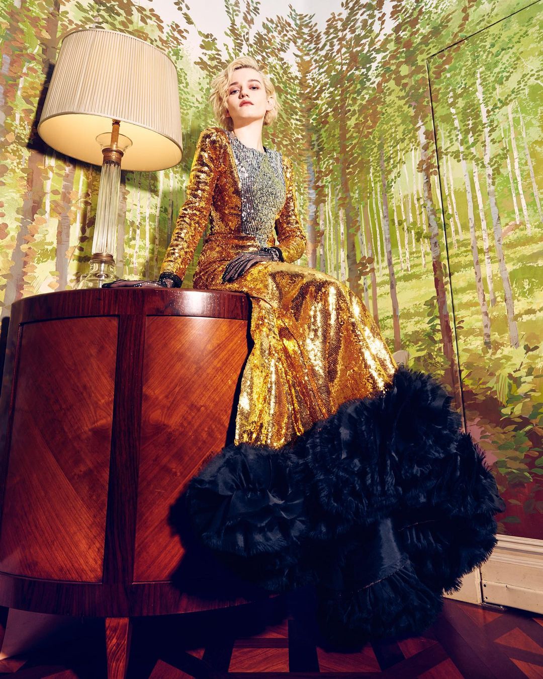 Julia Garner in a gold sequin gown, seated on a wood table with a lamp; forest-themed wallpaper backdrop, indoors; medium shot.