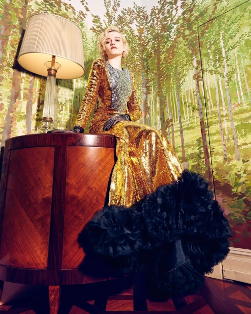 Julia Garner in a gold sequin gown, seated on a wood table with a lamp; forest-themed wallpaper backdrop, indoors; medium shot.