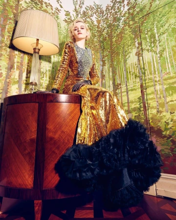 Julia Garner in a gold sequin gown, seated on a wood table with a lamp; forest-themed wallpaper backdrop, indoors; medium shot.