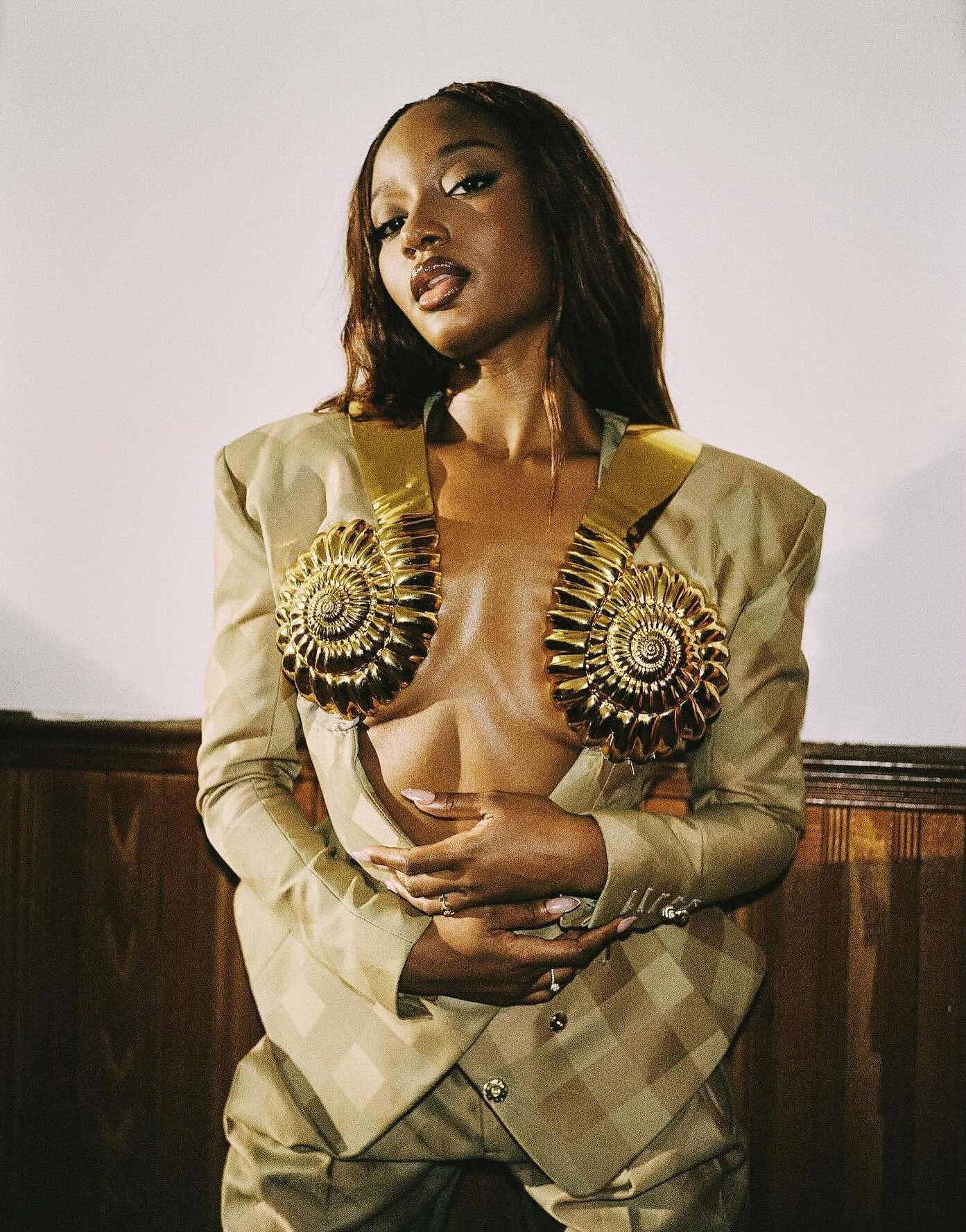 Portrait of a stylish Black woman in a gold and beige suit with nautilus shell details, posing indoors in front of a paneled wall.