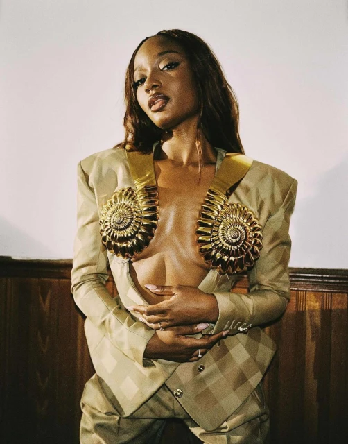 Portrait of a stylish Black woman in a gold and beige suit with nautilus shell details, posing indoors in front of a paneled wall.