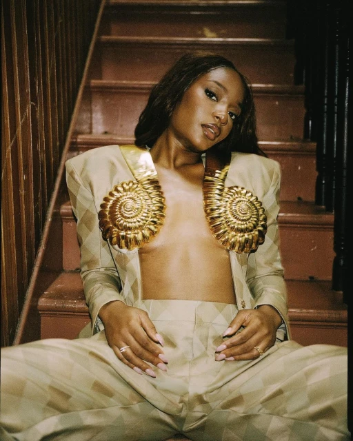 Portrait of an adult woman in a beige suit with golden shell details, sitting on wooden stairs with a neutral facial expression.
