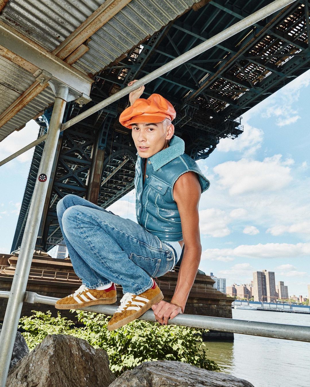 Stylish man squats on a metal rail wearing a shiny blue vest, jeans, a hat, and sneakers with a city bridge in the background.