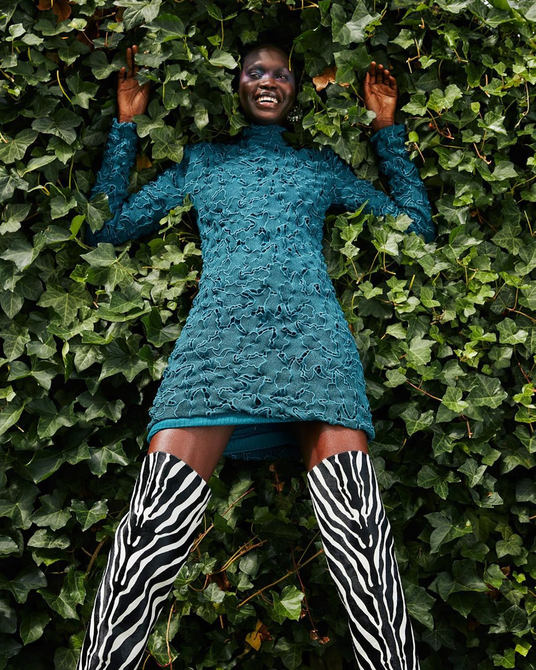 Smiling black woman in green dress and zebra boots lying on green ivy background, outdoor fashion shot, eye-level angle.