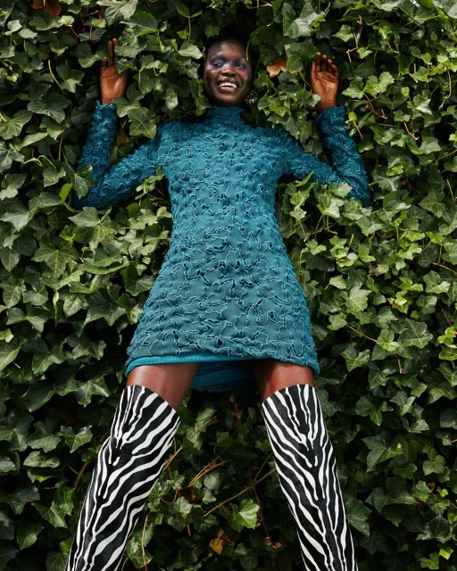 Smiling black woman in green dress and zebra boots lying on green ivy background, outdoor fashion shot, eye-level angle.