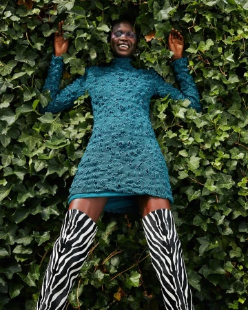 Smiling black woman in green dress and zebra boots lying on green ivy background, outdoor fashion shot, eye-level angle.