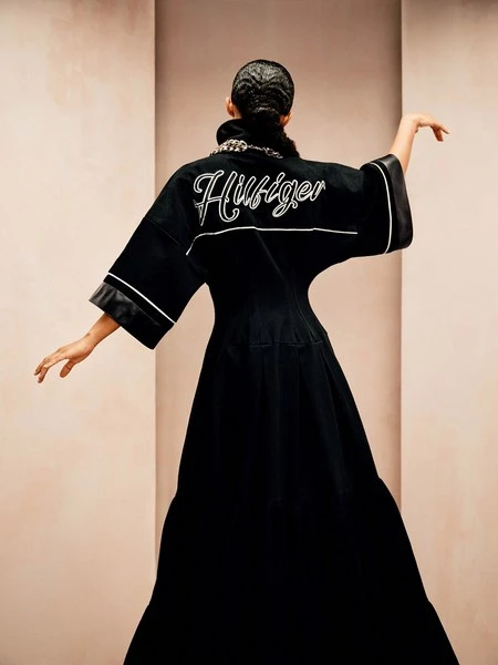 A woman in a floor-length black jacket with the word “Hilfiger” written in cursive across the back is posing with her arms outstretched.