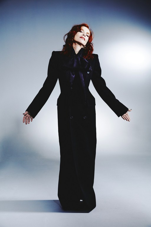 Elegant woman in a long black jacket and pants with hands outstretched in a photo studio setting, illuminated by soft, diffused lighting.