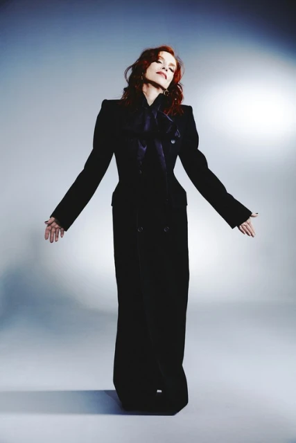 Elegant woman in a long black jacket and pants with hands outstretched in a photo studio setting, illuminated by soft, diffused lighting.