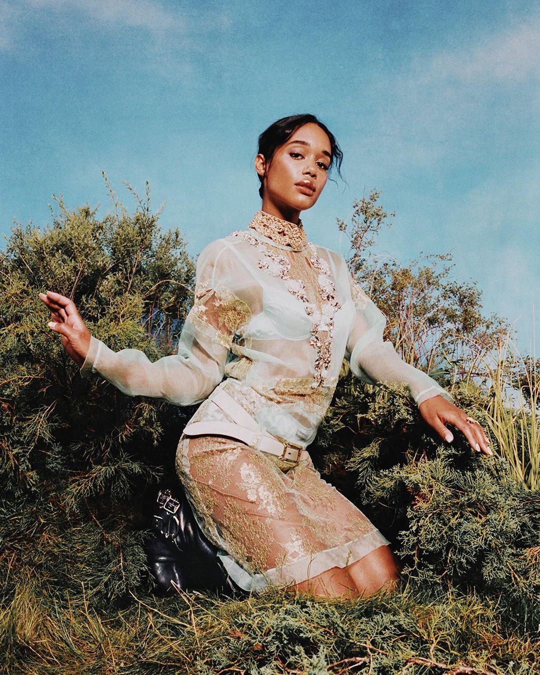 Laura Harrier models in a sheer lace dress, kneeling among greenery beneath a bright sky, striking a pose with her hands.