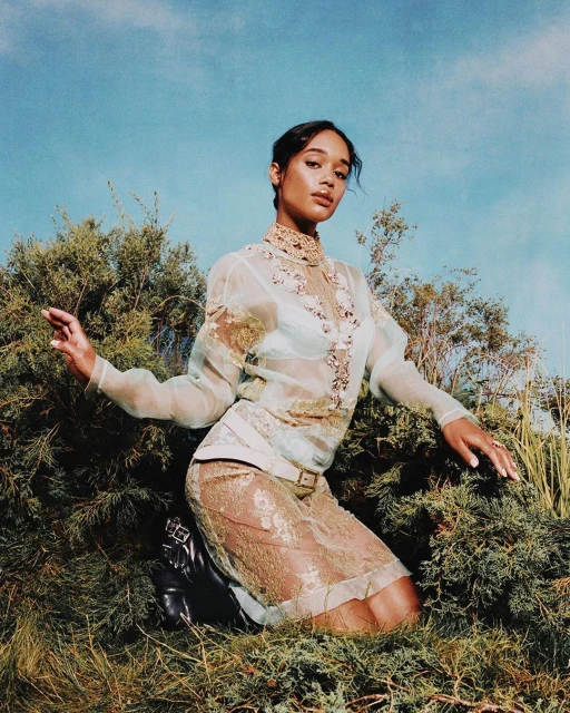 Laura Harrier models in a sheer lace dress, kneeling among greenery beneath a bright sky, striking a pose with her hands.
