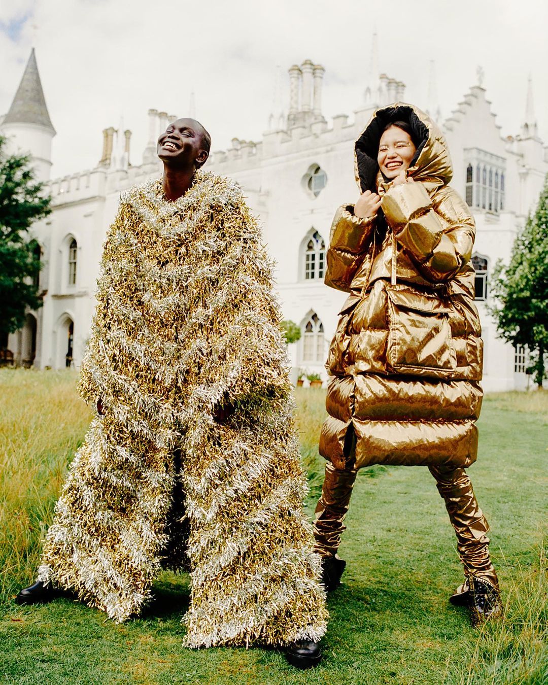 Two smiling fashion models pose outdoors against a castle backdrop, wearing unique, oversized metallic gold and fringe outfits.