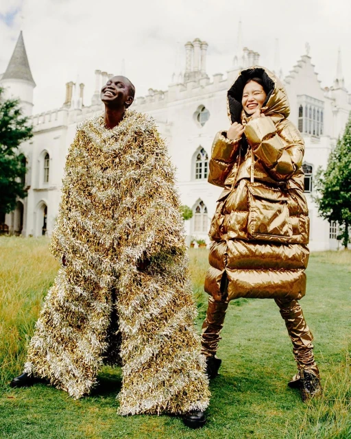 Two smiling fashion models pose outdoors against a castle backdrop, wearing unique, oversized metallic gold and fringe outfits.