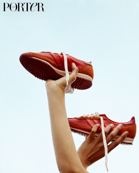 A pair of coral-red running shoes is held up against a clear sky by a person with outstretched arms. Porter Magazine logo visible.