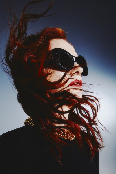 Red-haired woman with black sunglasses, wearing a gold chain, looking up with wind blowing through her hair against a gradient background.