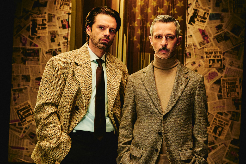 Two adult men stand in front of a backdrop of old newspapers, wearing vintage-style suits and looking directly at the camera.