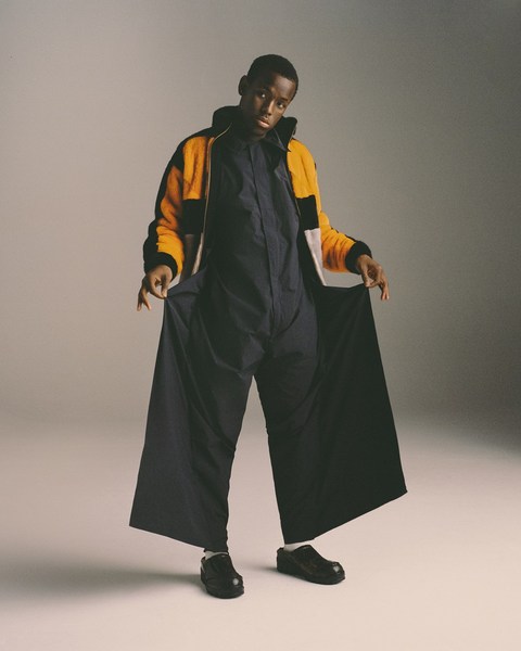 Full-length shot of an African-American male model in a navy jumpsuit and black shoes, posing in a light fur coat on a grey background.