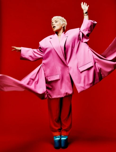 Stylish woman in oversized pink suit and orange pants strikes a pose against a red backdrop, showcasing a vibrant and fashionable look.