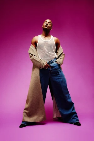 Black man wearing a white tank top, blue jeans, and an open tan coat against a vibrant purple backdrop.