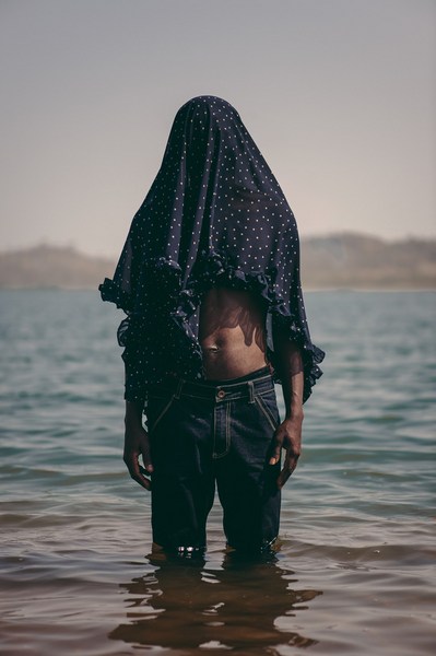 Figure standing waist-deep in water wearing a dark polka dot fabric draped over their head and dark denim jeans.
