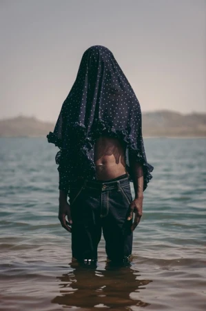 Figure standing waist-deep in water wearing a dark polka dot fabric draped over their head and dark denim jeans.