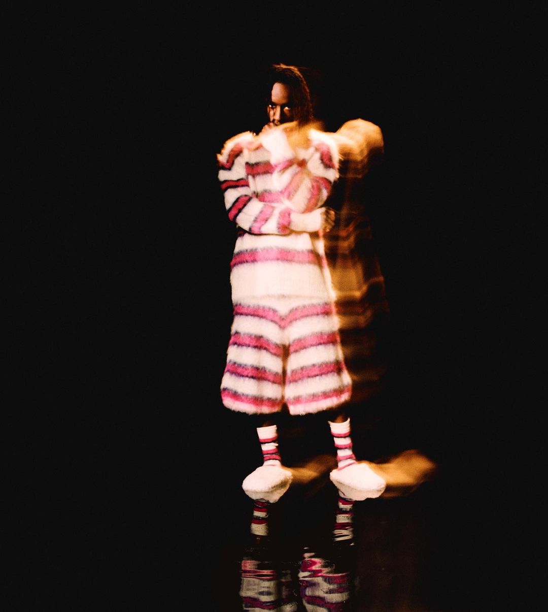 An adult with brown hair, wearing a horizontally striped pink and white fuzzy outfit, standing in a dimly lit, black-toned photo studio.