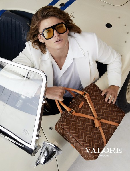 Masculine fashion portrait: Man in cream convertible, wearing sunglasses, holding a patterned duffel bag, showcasing style and luxury.