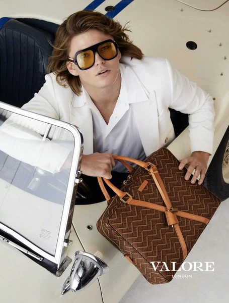 Masculine fashion portrait: Man in cream convertible, wearing sunglasses, holding a patterned duffel bag, showcasing style and luxury.