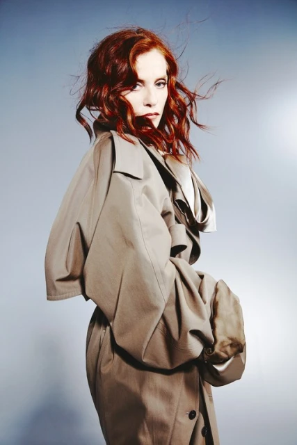 Portrait of a red-haired woman wearing a tan trench coat with a serious expression against a grey background in a studio setting.