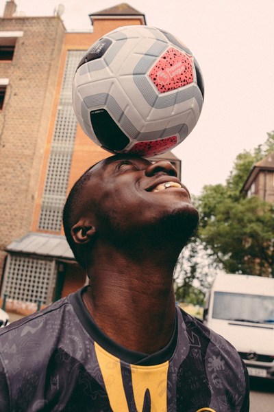 Smiling adult male balancing a soccer ball on his head in an outdoor setting with brick buildings and a van in the background.