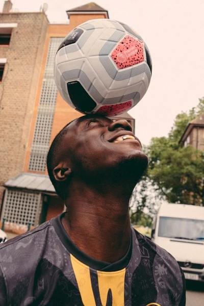 Smiling adult male balancing a soccer ball on his head in an outdoor setting with brick buildings and a van in the background.