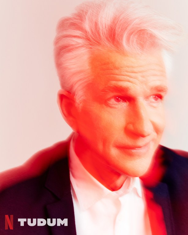 Headshot of adult male with silver hair, wearing a dark suit and white shirt, with a red filter effect and the text Netflix Tudum.